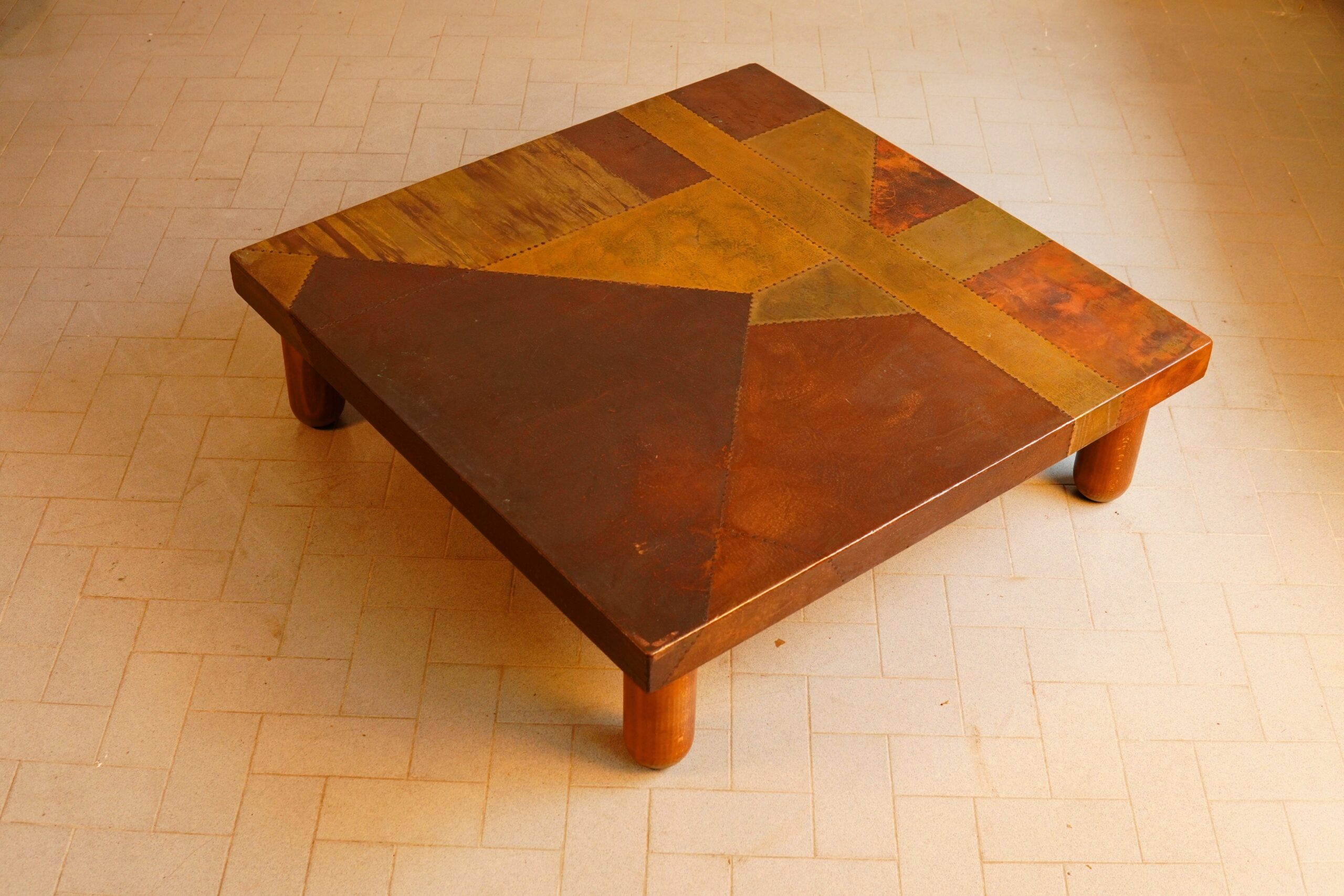 Lorenzo Burchiellaro (Venice, 1933 - 2017)An Important and Large Mid-Century Brutalist Coffee TableAtelier Burchiellaro, Italy, circa 1970.Description:A monumental and commanding low table of square proportions, epitomising the raw, sculptural elegance of the post-war Italian avant-garde while retaining a stately presence suited to the finest country house drawing room. The piece features four robust, cylindrical stained beech legs supporting a heavy, architectural top. The surface is meticulously clad in a geometric patchwork of hand-wrought copper plates, masterfully secured with exposed perimetric rivets. The metal displays an exceptional, undisturbed natural patination, yielding deep burnished tones, golden hues, and subtle iridescence born of half a century of gentle oxidation.Provenance:Private Collection.Condition Report:In very good country house condition. The timber structure remains structurally sound. The copper top retains its magnificent original patina with minor surface wear, light scratches, and intentional variances in tone consistent with age, authentic use, and the artist's original tactile intent.#LorenzoBurchiellaro#BrutalistDesign#AuctionHouse#QuietLuxury#FineFurniture